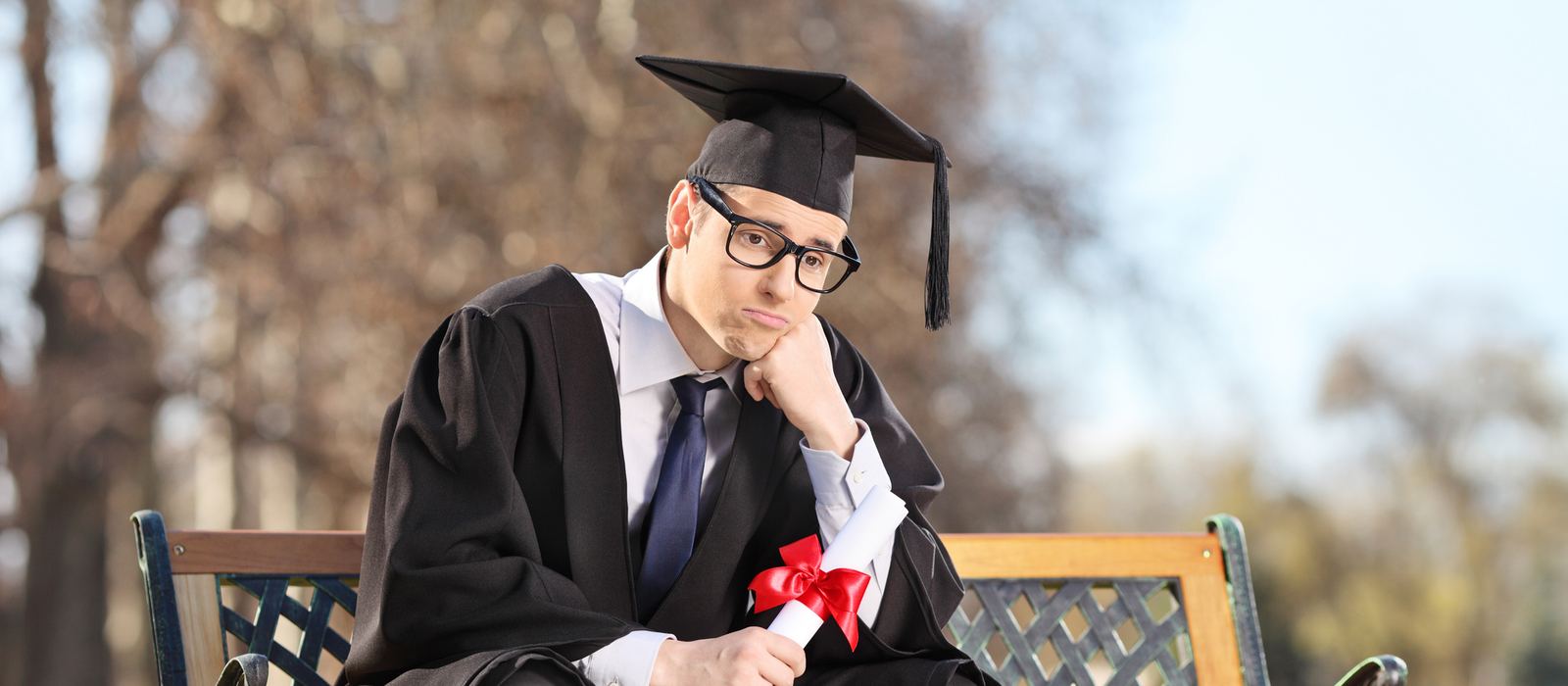 College graduate in cap and gown sitting alone on a park bench, holding his diploma and looking discouraged