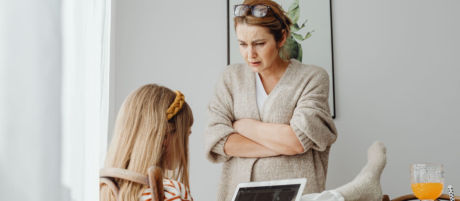 Mother standing with arms crossed, looking over her young adult daughter's shoulder as the daughter works on a laptop at home